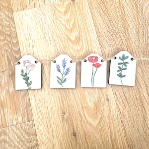 May include: A garland of five wooden tags with floral illustrations. Each tag features a different flower or plant design, including pink flowers, blue flowers, red flowers, and green leaves. The tags are connected by a twine string.