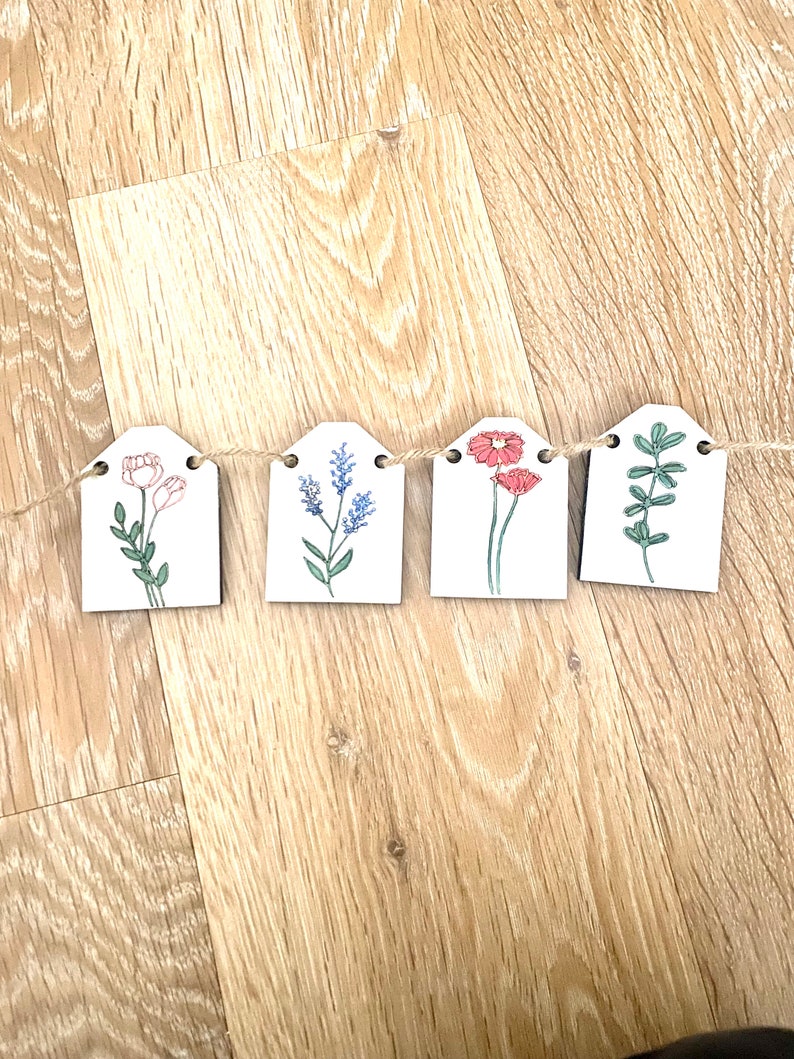 May include: A garland of five wooden tags with floral illustrations. Each tag features a different flower or plant design, including pink flowers, blue flowers, red flowers, and green leaves. The tags are connected by a twine string.