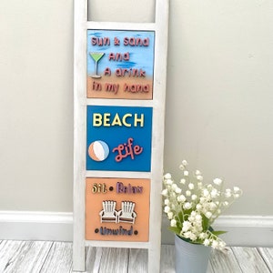 May include: A white wooden ladder with three signs. The top sign is blue and says "sun & sand And A drink in my hand". The middle sign is blue and says "BEACH Life". The bottom sign is orange and says "Sit. Relax. Unwind."
