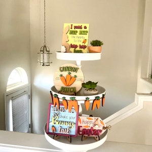 May include: A white tiered stand with three levels decorated with Easter-themed wooden signs. The top level features a sign that says "I said a hip hop the hippity to the hip hip hoo". The middle level has a sign that says "Carrot Patch" with a hand-picked and locally grown carrot design. The bottom level has two signs, one that says "Time to shake your bunny tail" and the other that says "Happy Easter".