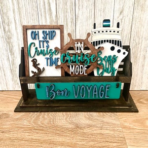 May include: Wooden sign holder with a teal sign that says "Bon Voyage" and other signs that say "Oh Ship It's Cruise Time", "In Cruise Mode", and "Seas The Day".