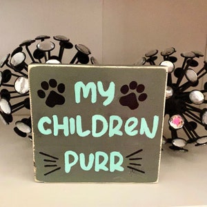 Op de afbeelding: Een grijs houten bord met zwarte poot afdrukken en de tekst "Mijn kinderen purren" in turquoise blauwe verf.