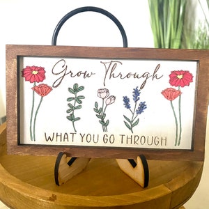May include: Wooden sign with a brown frame and a white background. The sign has a black metal handle and a floral design with the text "Grow Through What You Go Through".