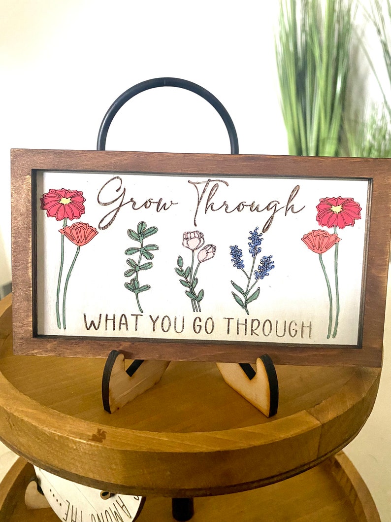 May include: Wooden sign with a brown frame and a white background. The sign has a black metal handle and a floral design with the text "Grow Through What You Go Through".