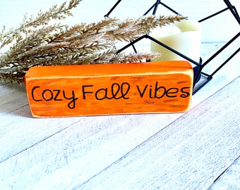 May include: Orange wooden sign with the text "Cozy Fall Vibes" written in black paint.