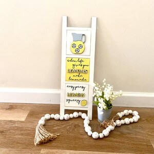 May include: White wooden ladder shelf with three yellow and white signs. The top sign has a yellow pitcher with ice cubes. The middle sign says "when life gives you lemons make lemonade". The bottom sign says "easy peasy lemon squeezy".