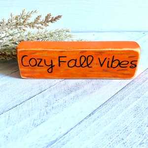 Pu&ograve; includere: Un cartello in legno arancione con la scritta "Cozy Fall Vibes" in vernice nera.
