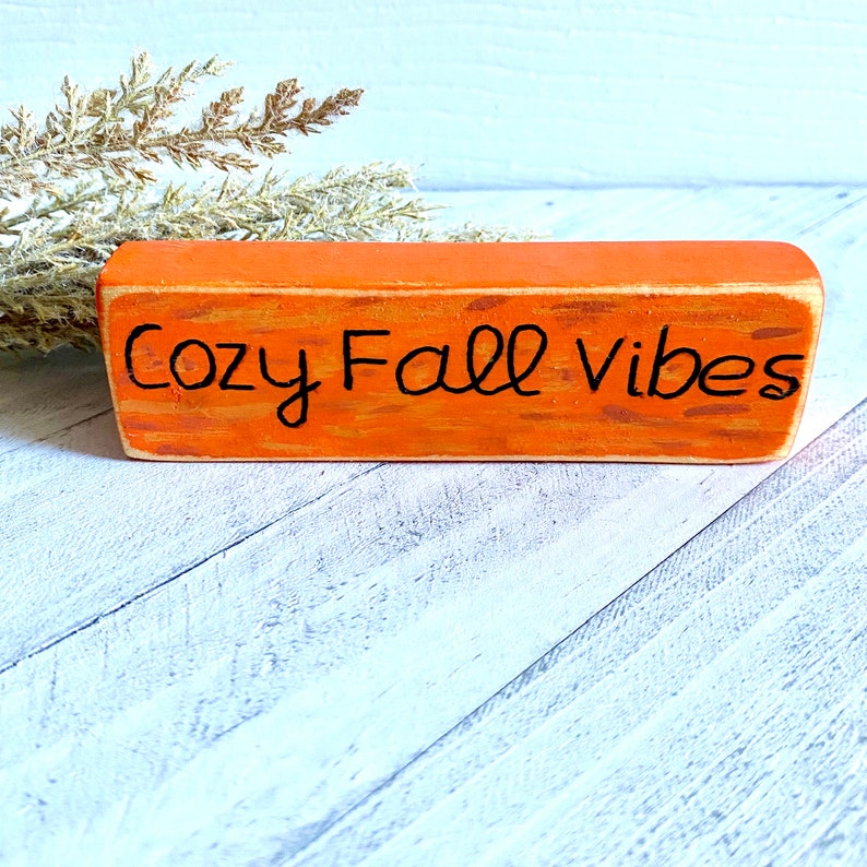 Pu&ograve; includere: Un cartello in legno arancione con la scritta "Cozy Fall Vibes" in vernice nera.