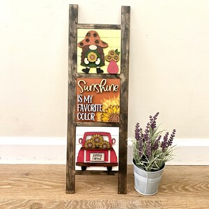 May include: A wooden ladder shelf with three signs. The top sign features a cartoon gnome with sunflowers. The middle sign says "Sunshine is my favourite colour" with a sunflower graphic. The bottom sign features a red truck with sunflowers in the back and the words "sunny farms".