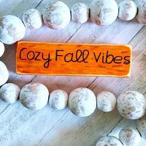 Pu&ograve; includere: Un cartello di legno con il testo "Cozy Fall Vibes" dipinto in nero su uno sfondo arancione. Il cartello &egrave; circondato da una ghirlanda di perline di legno bianche.