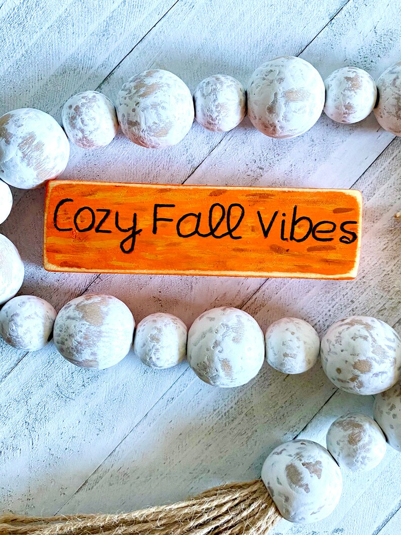 Pu&ograve; includere: Un cartello di legno con il testo "Cozy Fall Vibes" dipinto in nero su uno sfondo arancione. Il cartello &egrave; circondato da una ghirlanda di perline di legno bianche.