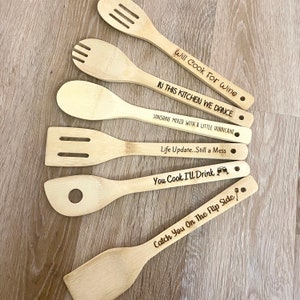 May include: Set of five wooden kitchen spoons with engraved sayings. The sayings include "Will Cook For Wine", "In This Kitchen We Dance", "Sunshine Mixed With A Little Hurricane", "Life Update...Still A Mess", and "You Cook I'll Drink".