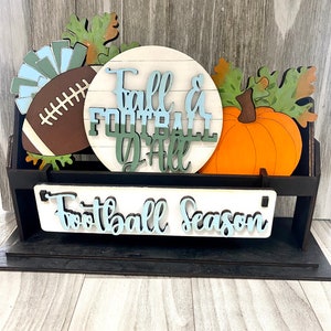 May include: A wooden sign with the text "Fall & Football Y'all" in blue and green lettering. The sign is decorated with a football, a pumpkin, and autumn leaves. The sign also has a smaller sign below it that says "Football Season".