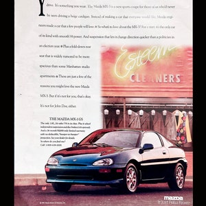 May include: A dark green Mazda MX-3 GS sports car is parked in front of a storefront with a sign that reads "Esteem Cleaners". The car has a black bumper and black wheels. The text "Mazda It Just Feels Right" is printed in white on a black background at the bottom of the image.