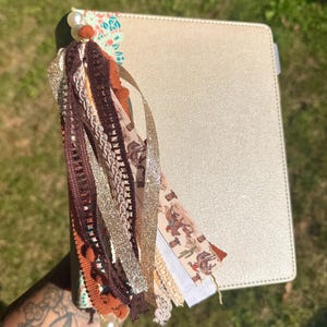 May include: A cream-colored journal featuring a decorative tassel made of various ribbons, including brown lace, gold glitter, and a patterned ribbon with cacti and cowboy boots. The tassel is accented with white pearl beads and small orange pom-poms.