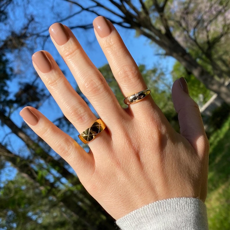 May include: Two gold rings worn on fingers. One ring is a wide band featuring a raised, crisscross quilted pattern. The second ring is a thinner, rounded band decorated with small, engraved star shapes across its polished surface.