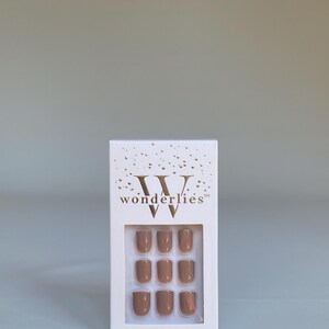 May include: A white rectangular box containing nine press-on nails in a solid tan color. The packaging features gold foil confetti accents at the top, the brand name "wonderlies" in gold lettering, and the text "MINUTE MANI" at the bottom.