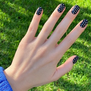 May include: A hand with fingernails painted in a glossy black polish featuring a white polka dot pattern. The nails are shaped with rounded tips, and the dark base color contrasts with the small, evenly spaced white dots across each nail surface.