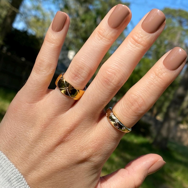 May include: Two gold-toned rings worn on fingers with light brown nail polish. One ring features a raised, quilted diamond pattern across its surface. The second ring is a smooth, rounded band accented with small, embedded star-shaped details.