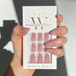 May include: A set of nude-colored press-on nails in a white box with the text "Wonderlies" and "Minute Mani" printed on it. The nails are arranged in rows of three, and the box is held by a hand.
