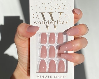 May include: A set of nude-colored press-on nails in a white box with the text "Wonderlies" and "Minute Mani" printed on it. The nails are arranged in rows of three, and the box is held by a hand.