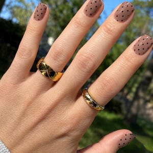 May include: Two gold rings worn on fingers with light brown nail polish featuring black polka dots. One ring has a wide, polished, quilted-style band, while the other is a thinner, rounded band with small, embedded, clear, sparkling stones.