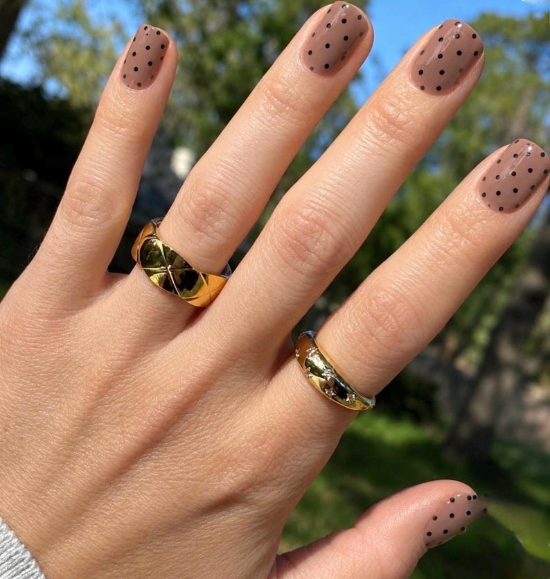 May include: Two gold rings worn on fingers with light brown nail polish featuring black polka dots. One ring has a wide, polished, quilted-style band, while the other is a thinner, rounded band with small, embedded, clear, sparkling stones.