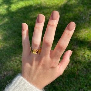 May include: A hand with nude nail polish is wearing a gold ring with a geometric design. The ring is on the middle finger.