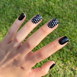 May include: Fingernails featuring a glossy finish with a combination of solid black polish and black polish with white polka dots. The index and pinky fingers are painted solid black, while the middle and ring fingers display a white polka dot pattern.