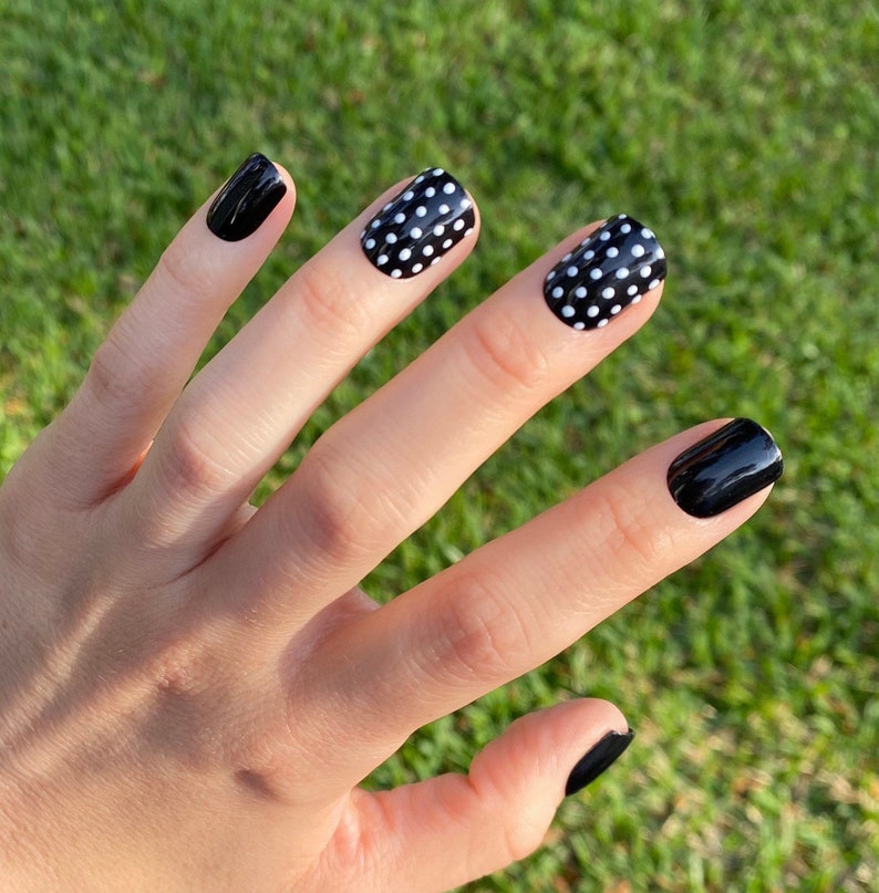 May include: Fingernails featuring a glossy finish with a combination of solid black polish and black polish with white polka dots. The index and pinky fingers are painted solid black, while the middle and ring fingers display a white polka dot pattern.