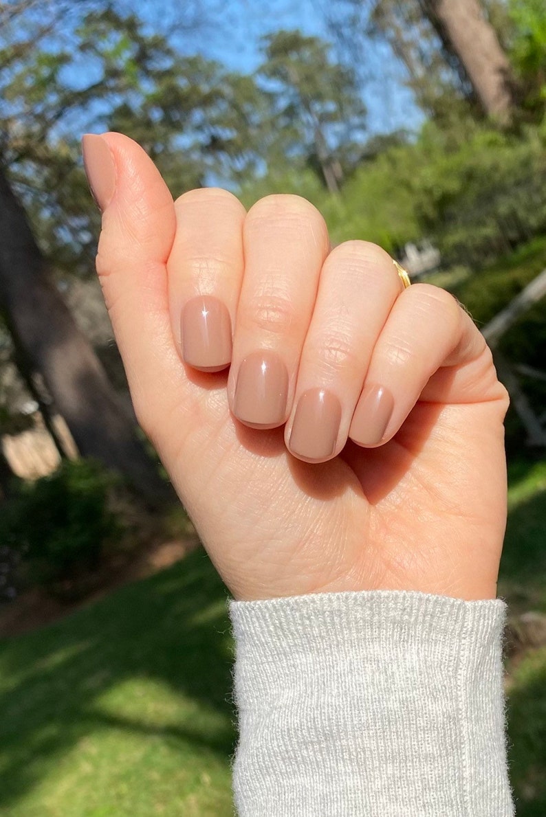 May include: A hand with short, rounded fingernails painted in a glossy, solid light taupe or beige nail polish. The nails have a smooth, reflective finish and are neatly groomed. The hand is positioned against a blurred outdoor background.
