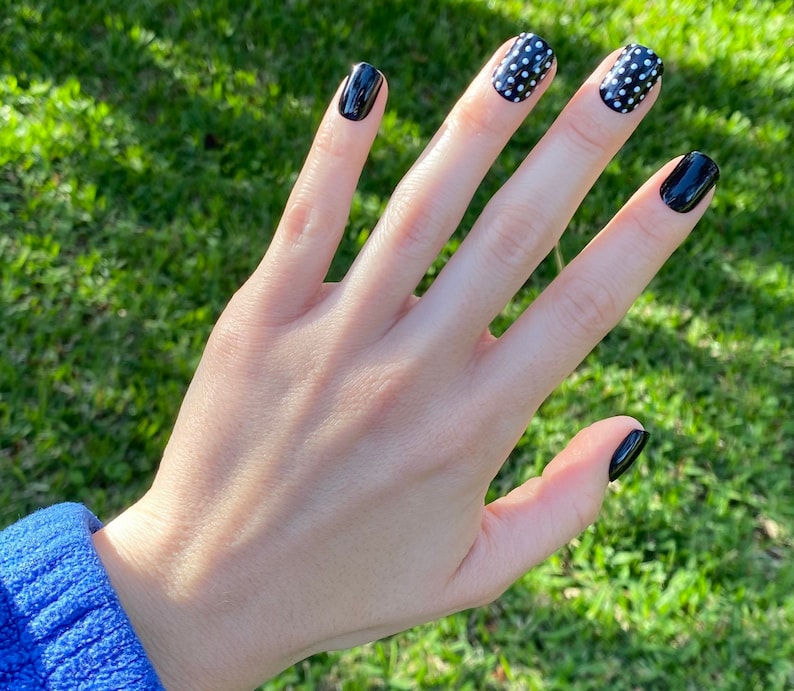 May include: Fingernails featuring a glossy finish with a combination of solid black polish and black polish with white polka dots. The nails are shaped with rounded edges, displaying a high-shine appearance across all five fingers on the hand.