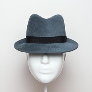 May include: A gray fedora hat with a black band. The hat is displayed on a white mannequin head against a white background. The hat has a classic, slightly indented crown and a brim that curves up slightly.