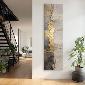 May include: Vertical abstract artwork with gold leaf and grey and white tones. The piece features a gold leaf design with black vein-like lines. It is displayed on a white wall in a modern interior setting.