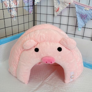 Puede incluir: Una cama para mascotas con forma de cerdo rosa de peluche, con ojos negros y un hocico rosa. La cama tiene una forma redondeada con una abertura en la parte delantera. El fondo incluye una rejilla negra y banderas decorativas coloridas.