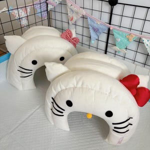 Puede incluir: Dos almohadas blancas con forma de gato con ojos y bigotes negros. Una tiene un lazo rojo y blanco a cuadros, y la otra un lazo rojo. Las almohadas están sobre una superficie blanca, con una pancarta decorativa en el fondo.