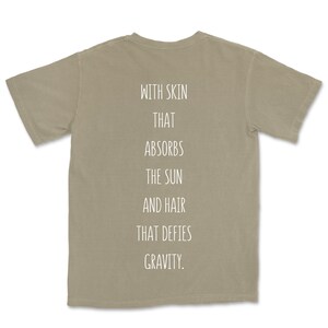 May include: A light brown t-shirt with the text "WITH SKIN THAT ABSORBS THE SUN AND HAIR THAT DEFIES GRAVITY." printed on the back.