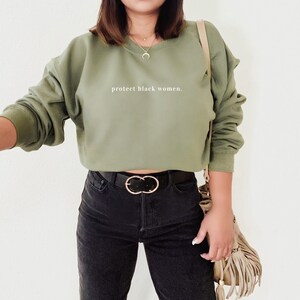 May include: A green cropped sweatshirt with the text "protect black women." The sweatshirt is worn with black jeans and a brown belt with a gold buckle.