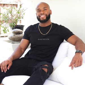K&ouml;nnte beinhalten: Ein Mann tr&auml;gt ein schwarzes T-Shirt mit dem Aufdruck "JUNETEENTH." Er sitzt auf einem wei&szlig;en Sofa und tr&auml;gt eine goldene Halskette und eine schwarze Uhr.