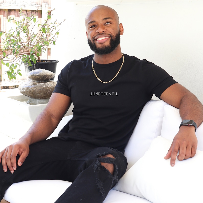 K&ouml;nnte beinhalten: Ein Mann tr&auml;gt ein schwarzes T-Shirt mit dem Aufdruck "JUNETEENTH." Er sitzt auf einem wei&szlig;en Sofa und tr&auml;gt eine goldene Halskette und eine schwarze Uhr.