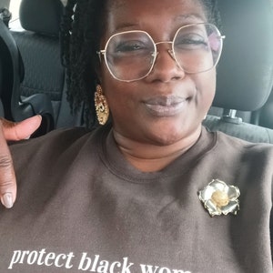 May include: A person wearing a brown sweatshirt with the text "protect black women." The sweatshirt has a gold flower pin on the left side.