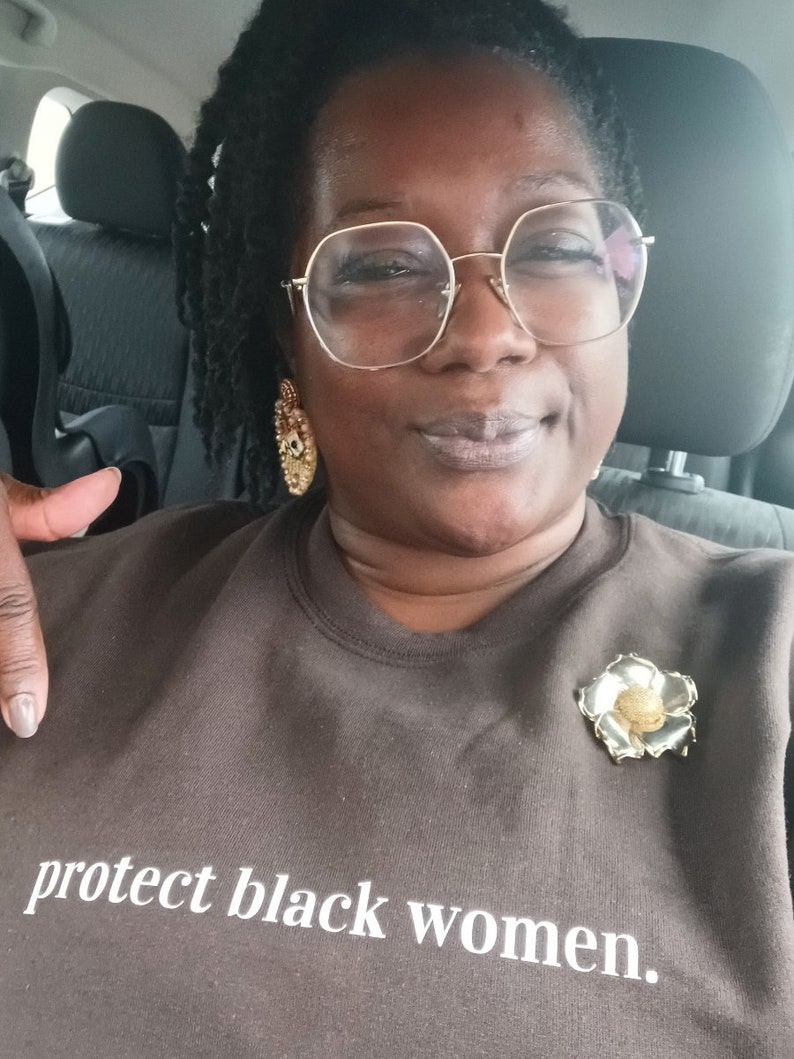 May include: A person wearing a brown sweatshirt with the text "protect black women." The sweatshirt has a gold flower pin on the left side.