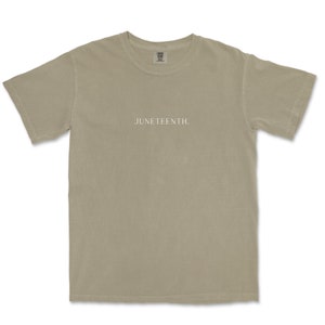 K&ouml;nnte beinhalten: Ein hellbeiges T-Shirt mit dem Aufdruck "JUNETEENTH." in Wei&szlig; auf der Vorderseite.