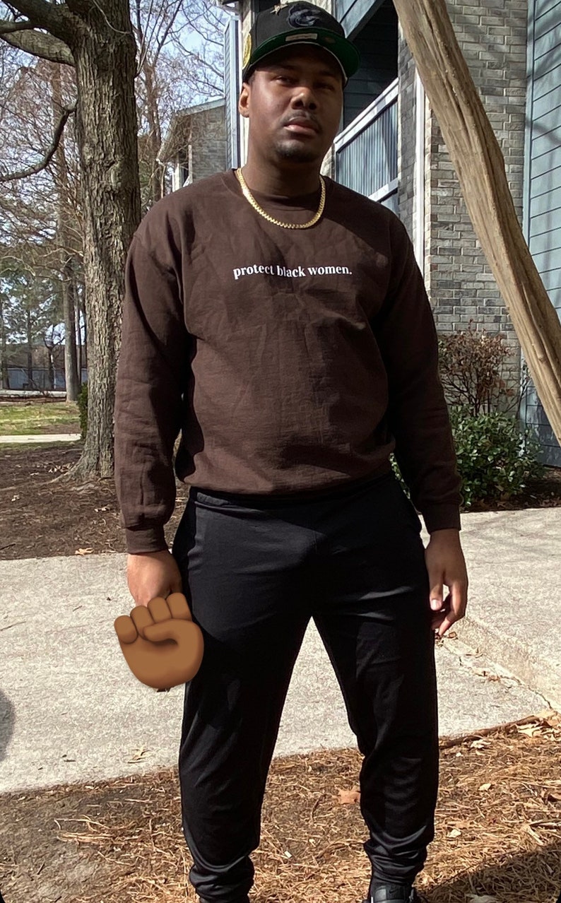 May include: A person wearing a brown long-sleeved shirt with the text "protect black women." printed on the front. The person is also wearing black pants and a black and green baseball cap.