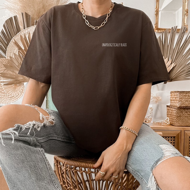 May include: A brown oversized t-shirt with the text "Unapologetically Black" printed on the front. The person is wearing the shirt with ripped denim jeans and a gold chain necklace.