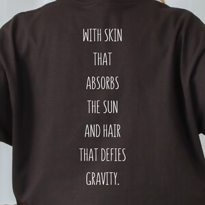 May include: Black t-shirt with white text that reads "With skin that absorbs the sun and hair that defies gravity."