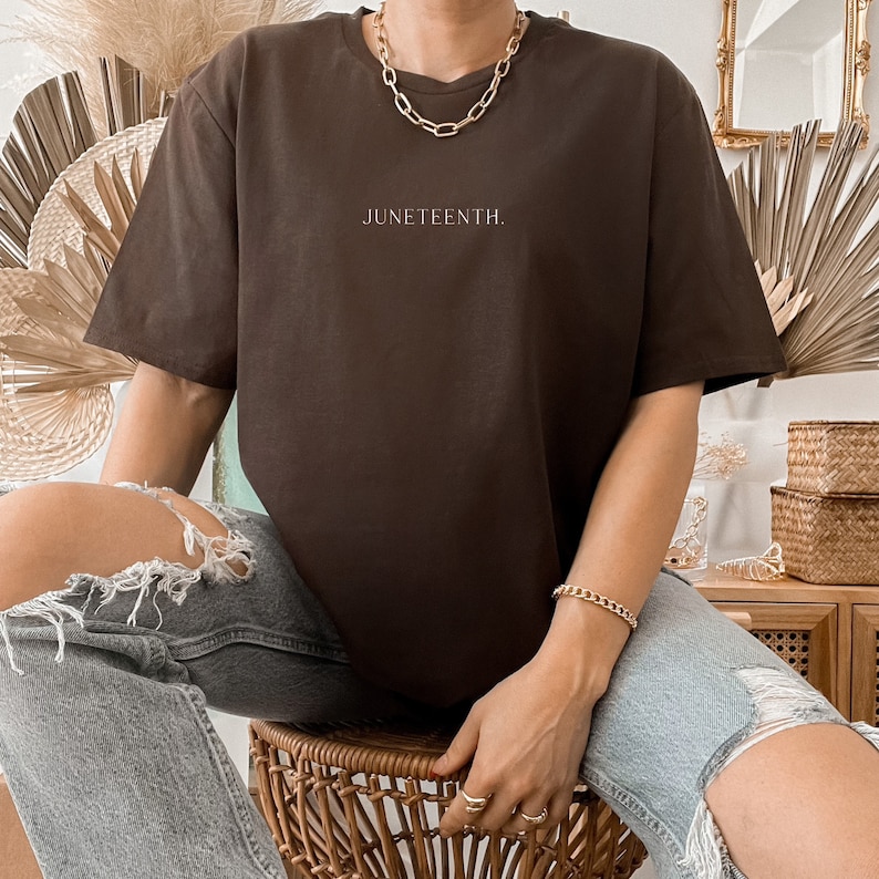 K&ouml;nnte beinhalten: Ein braunes T-Shirt mit dem Aufdruck "Juneteenth." auf der Vorderseite. Das Shirt wird von einer Person mit einer goldenen Kettenkette und einem goldenen Armband getragen.