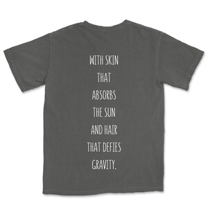 May include: A charcoal grey t-shirt with the text "WITH SKIN THAT ABSORBS THE SUN AND HAIR THAT DEFIES GRAVITY." printed on the back.