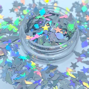 May include: A clear jar filled with iridescent holographic glitter shaped like stars, unicorns, and other whimsical shapes. The glitter is scattered around the jar on a white surface.