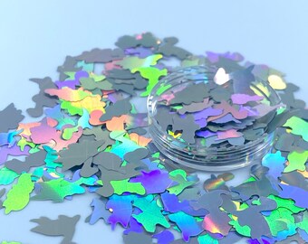 May include: A container of iridescent holographic dinosaur-shaped glitter. The glitter is a mix of green, blue, purple, and silver.